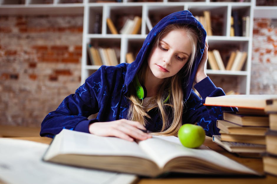 Teenage girl in blue hood reads in library, headphones and apple by her side
